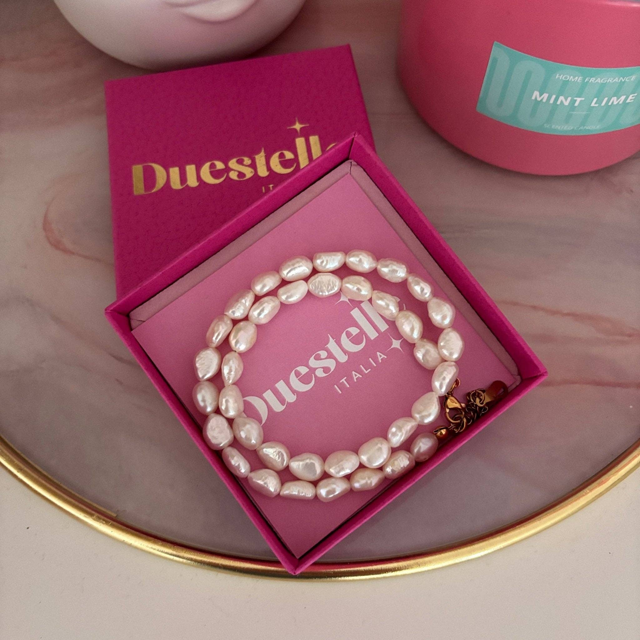 Pearl bracelet in a pink 'Duestell' box on a reflective surface with a pink container in the background.